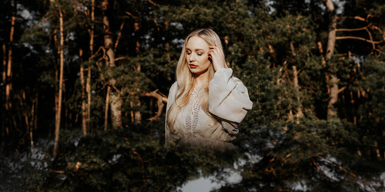 Eine blonde Frau steht im Mittelpunkt des Bildes, schaut nach unten und streicht mit ihrer linken Hand ihr Haar aus dem Gesicht. Im Hintergrund ist ein Wald zu sehen. In der unteren Hälfte ist der Wald gespiegelt zu sehen.
