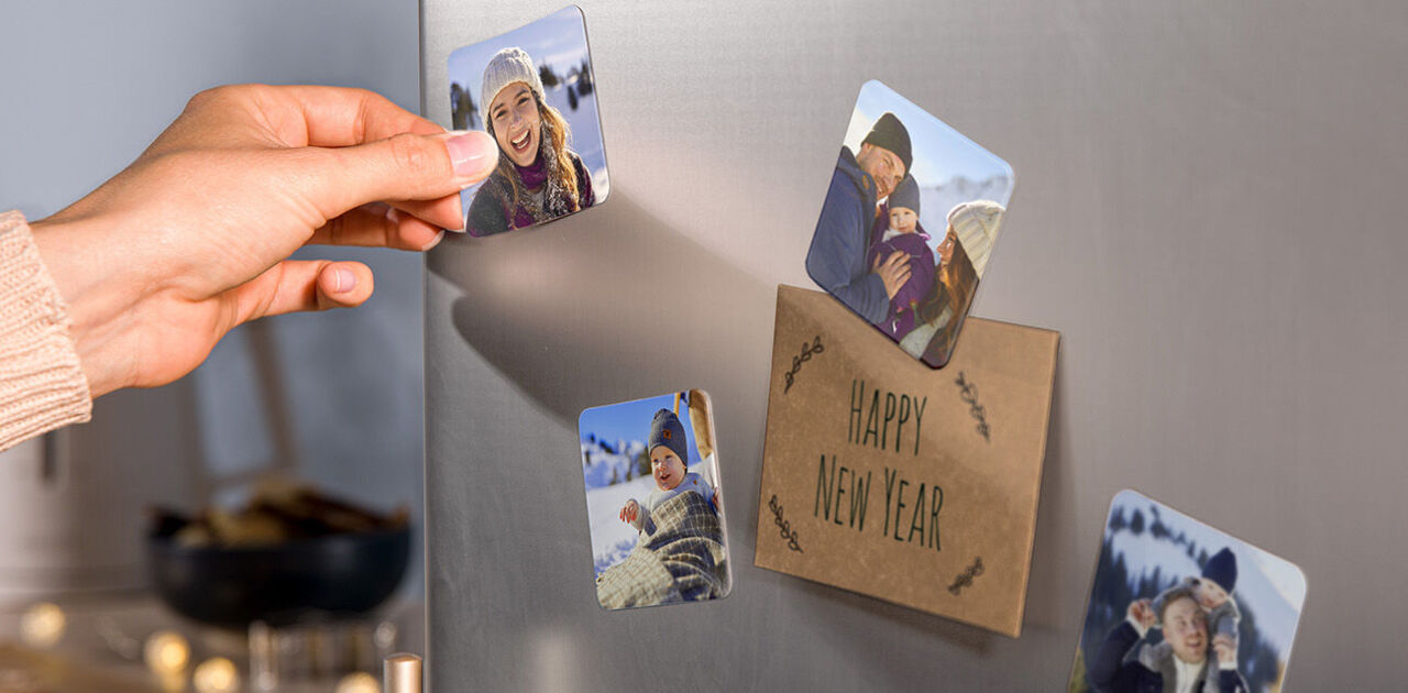 Person befestigt personalisierte Fotomagnete mit winterlichen Familienfotos an einem Kühlschrank neben einer Karte mit der Aufschrift „Happy