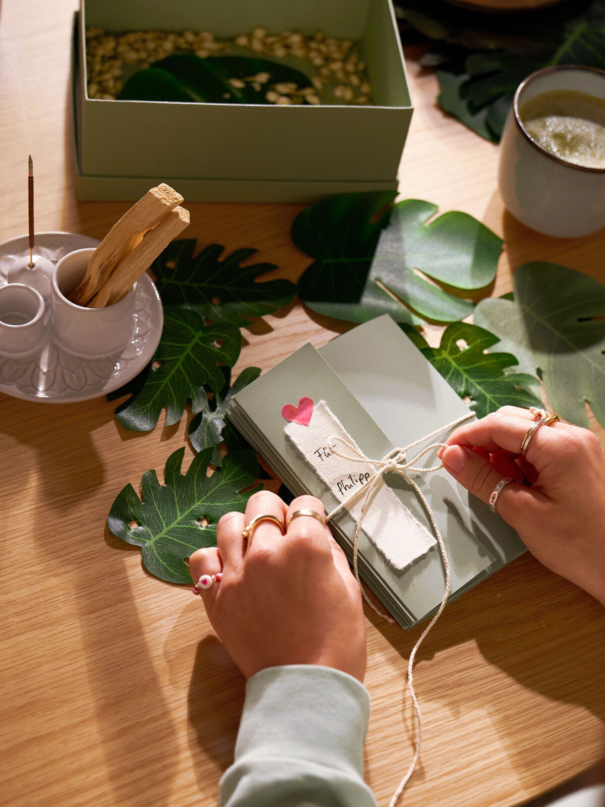 Auf einem Tisch ist eine Geschenkbox und Dekoration zu sehen. Eine Frau bindet eine Schleife um ein verpacktes Geschenk.