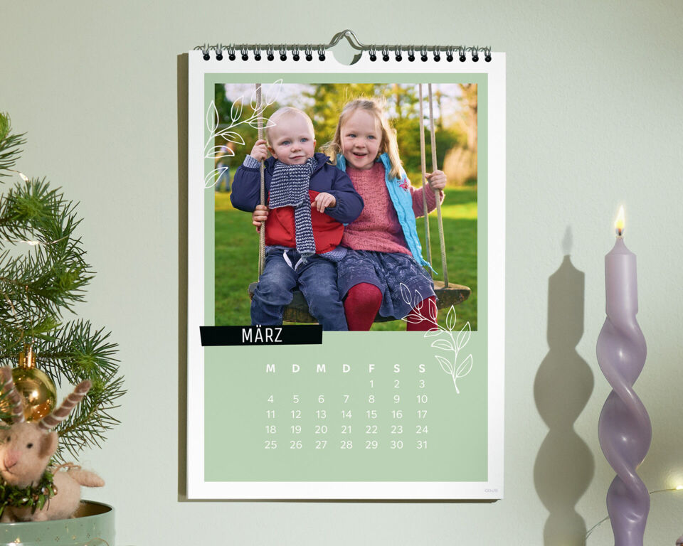 Personalisierter CEWE Fotokalender an der Wand mit einem Bild von zwei Kindern auf einer Schaukel, daneben eine Kerze und Weihnachtsdekorati