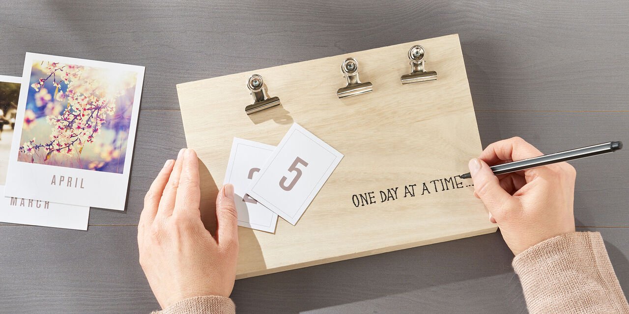 Zwei Hände schreiben den Satz "One day at a time…" auf die rechte Seite des Tischkalenders.