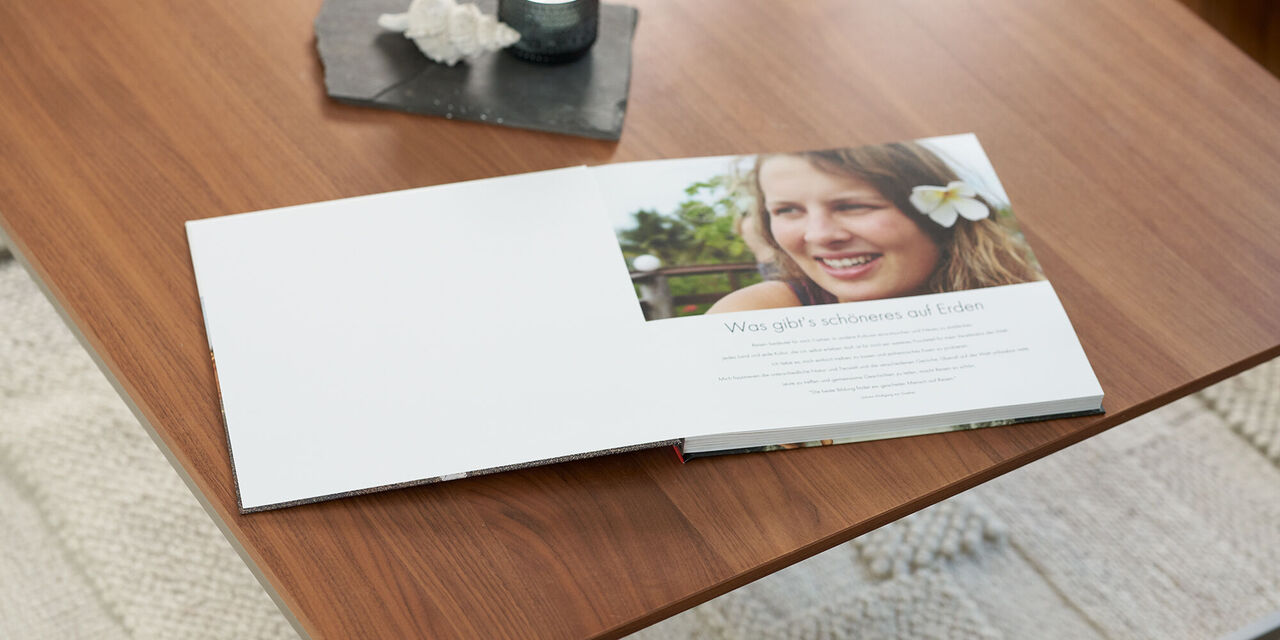 Auf einem Holztisch liegt ein aufgeschlagenes Fotobuch mit einem Einleitungstext und einem Foto von Lea Wippermann mit Blume im Haar. Über dem Fotobuch steht ein dunkles Tablett mit einer Kerze und Muschel.